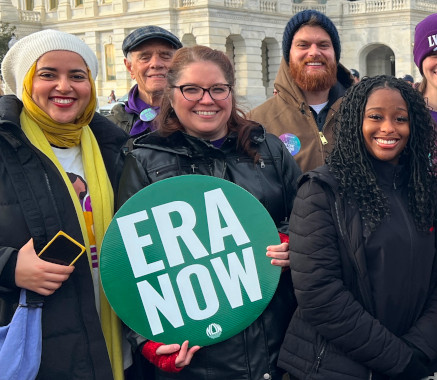 A smiling group of men and women, the lady in the center holds an 'ERA NOW' placard