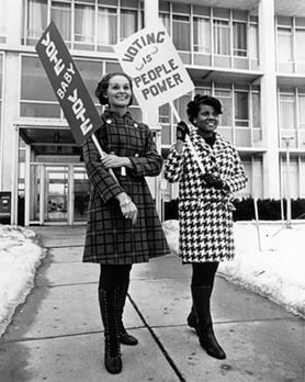 Two 1960s-era young ladies in stylish winter coats carry what look like protest signs: one sign says 'Vote, Baby, Vote' and the other says, 'Voting is People Power.'