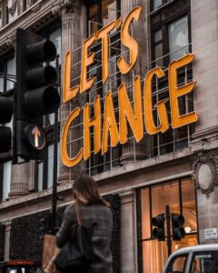 A person walks past a building with large illuminated yellow letters reading "LET'S CHANGE" above the entrance. Traffic lights and street signs are visible in the foreground.