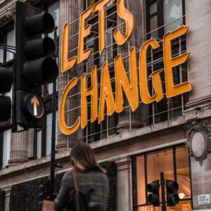 A person walks past a building with large illuminated yellow letters reading "LET'S CHANGE" above the entrance. Traffic lights and street signs are visible in the foreground.