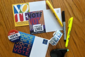Postcards, markers, pens, a roll of stamps, a correction tape, a “Your Vote Counts” button, and a highlighter arranged on a wooden surface, promoting voting and civic participation.