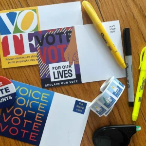 Postcards, markers, pens, a roll of stamps, a correction tape, a “Your Vote Counts” button, and a highlighter arranged on a wooden surface, promoting voting and civic participation.
